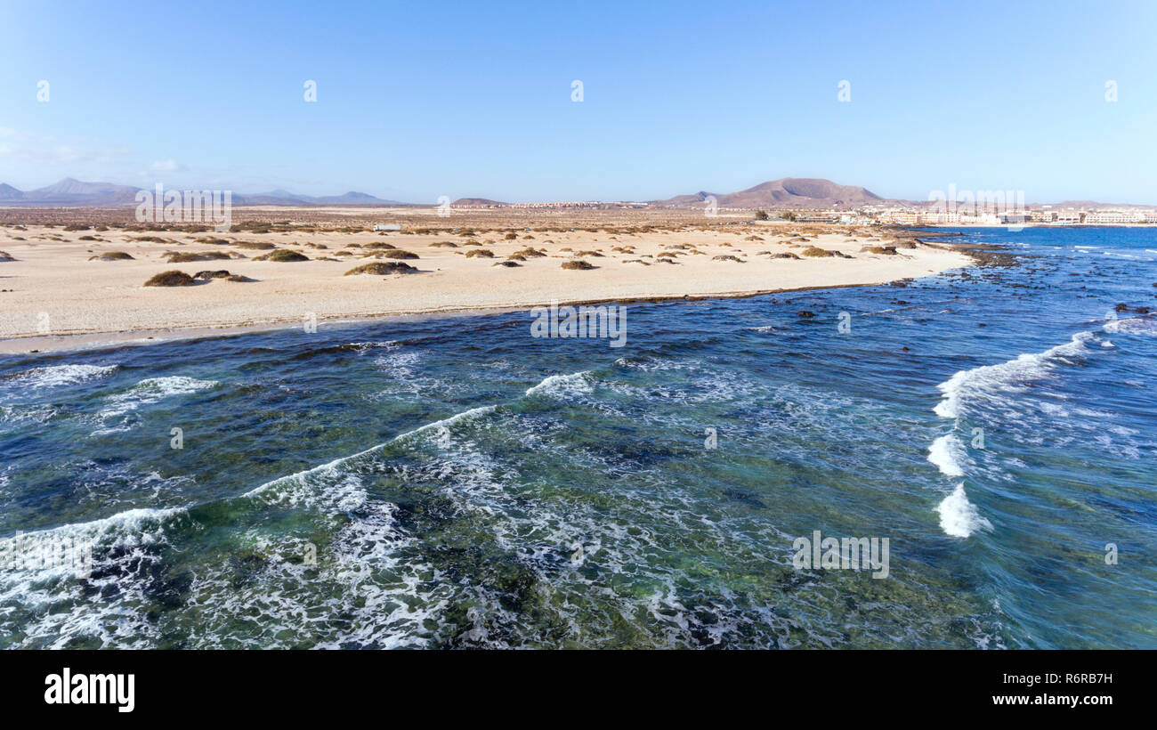 Aerial view of sea waves crushing white sand beach by dunes and volcanic mountains, on a sunny summer day in Fuerteventura, Canary Islands . Stock Photo