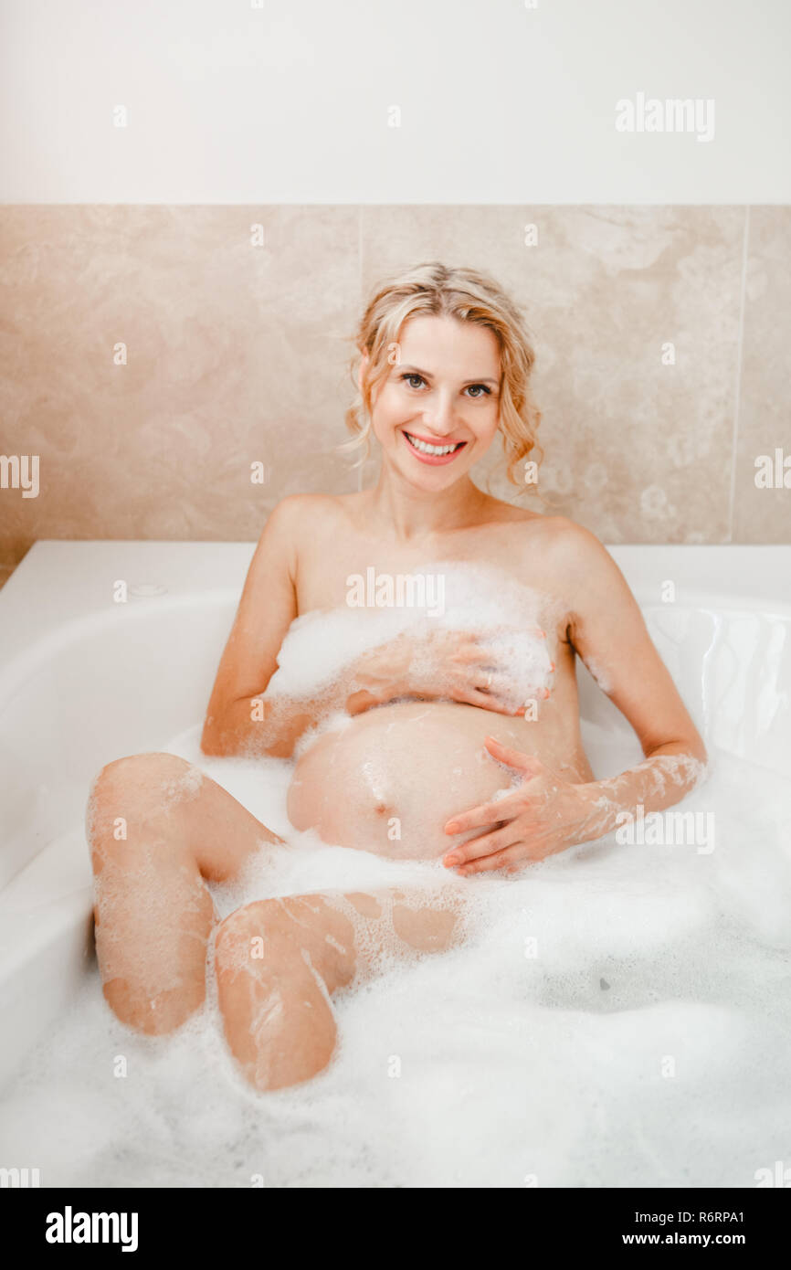 Happy healthy pregnancy. Portrait of pregnant blonde Caucasian woman washing in bathroom. Beautiful expecting mom lady taking morning bath with foam a Stock Photo