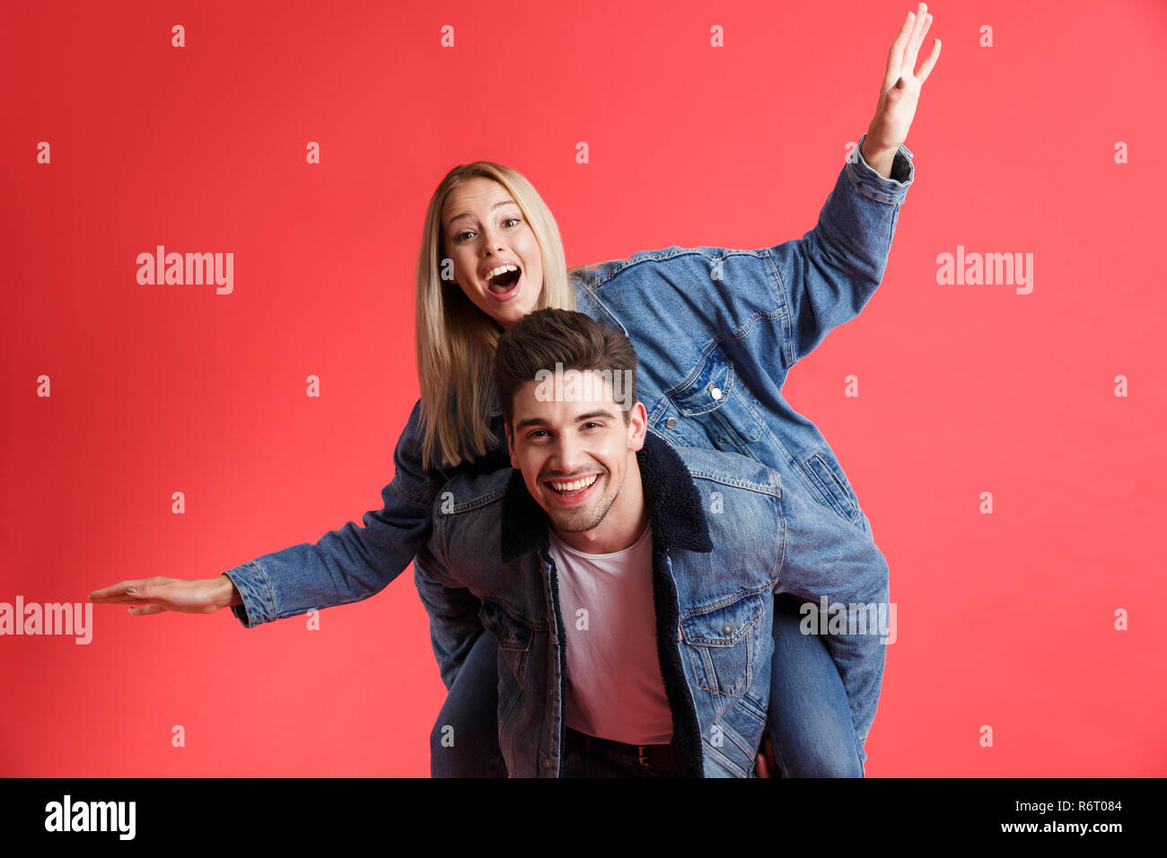 Portrait of a happy young couple dressed in denim jackets standing together isolated over red background, piggyback ride Stock Photo