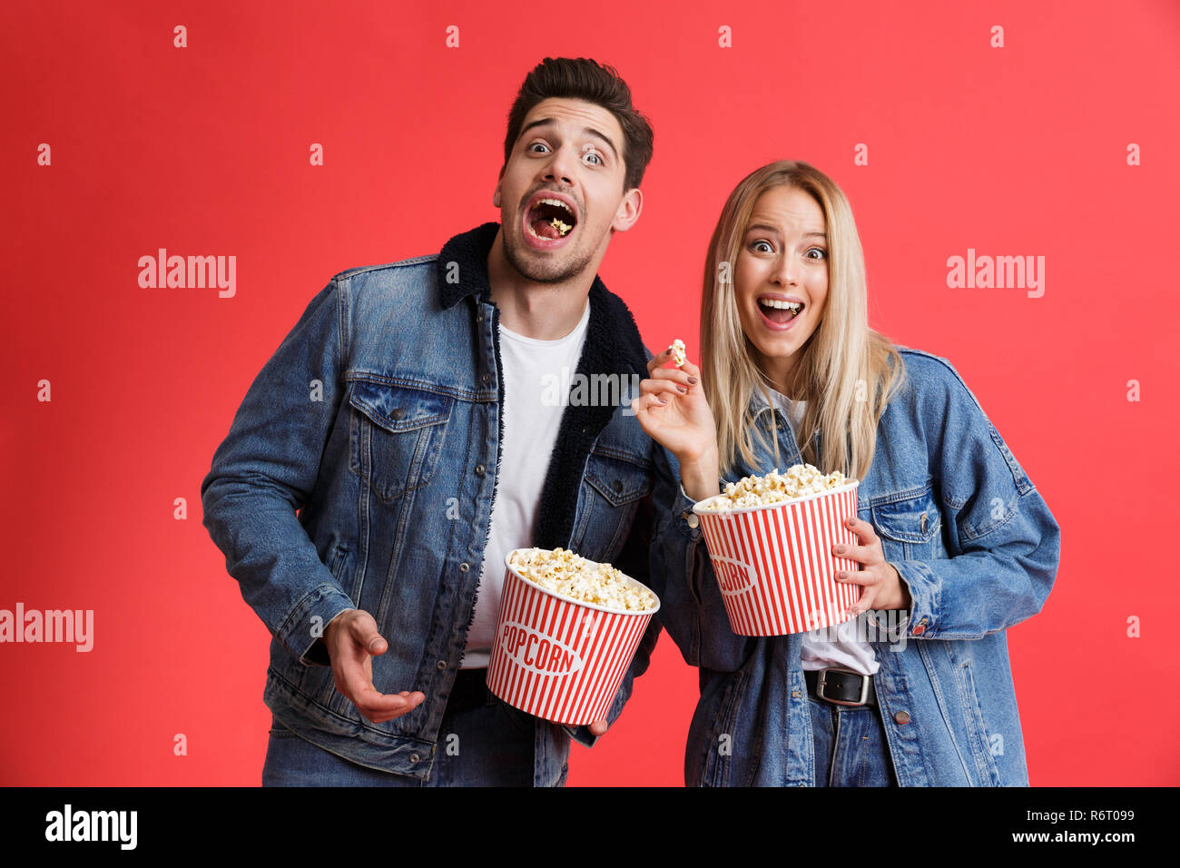 Portrait of an excited young couple dressed in denim jackets standing together isolated over red background, watching movie, eating popcorn Stock Photo