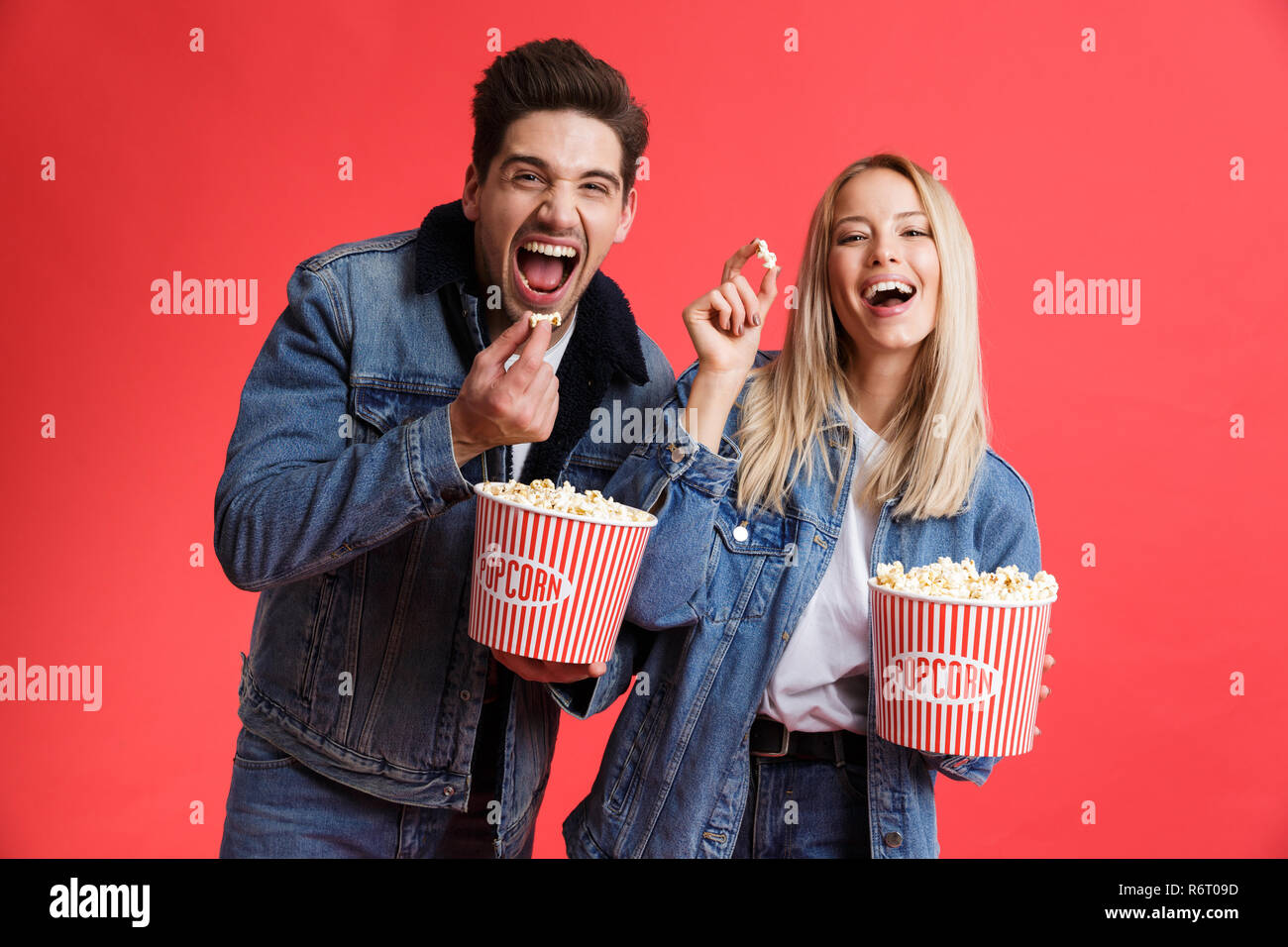 Portrait of a cheerful young couple dressed in denim jackets standing together isolated over red background, watching movie, eating popcorn Stock Photo