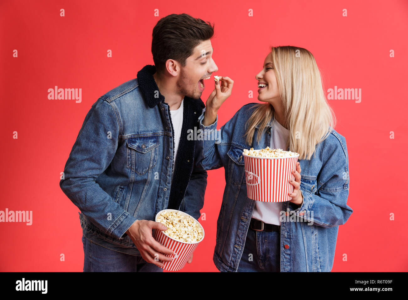 Portrait of a loving young couple dressed in denim jackets standing together isolated over red background, watching movie, eating popcorn Stock Photo