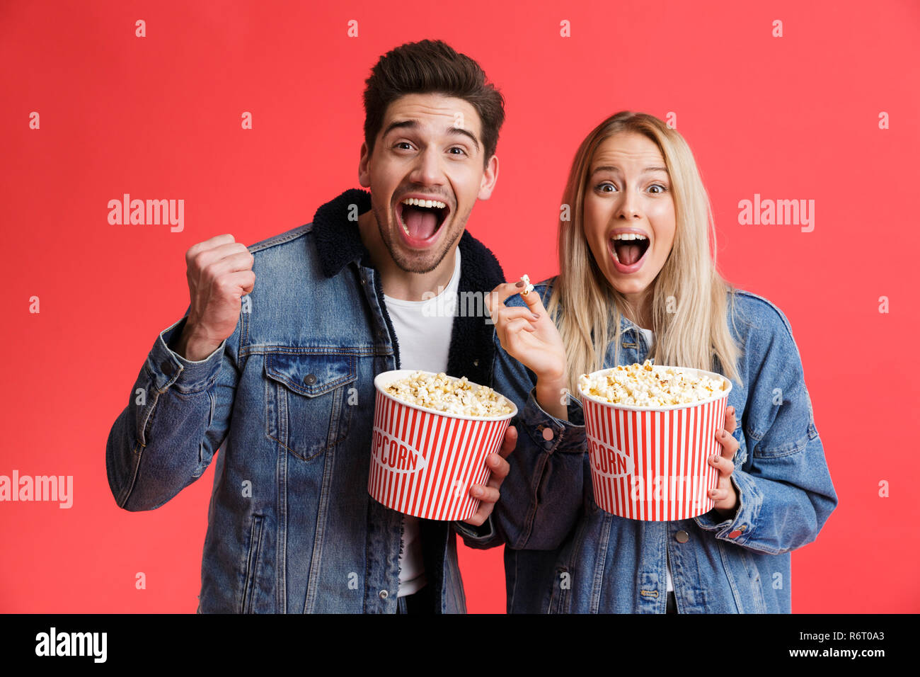 Portrait of an excited young couple dressed in denim jackets standing together isolated over red background, watching movie, eating popcorn Stock Photo
