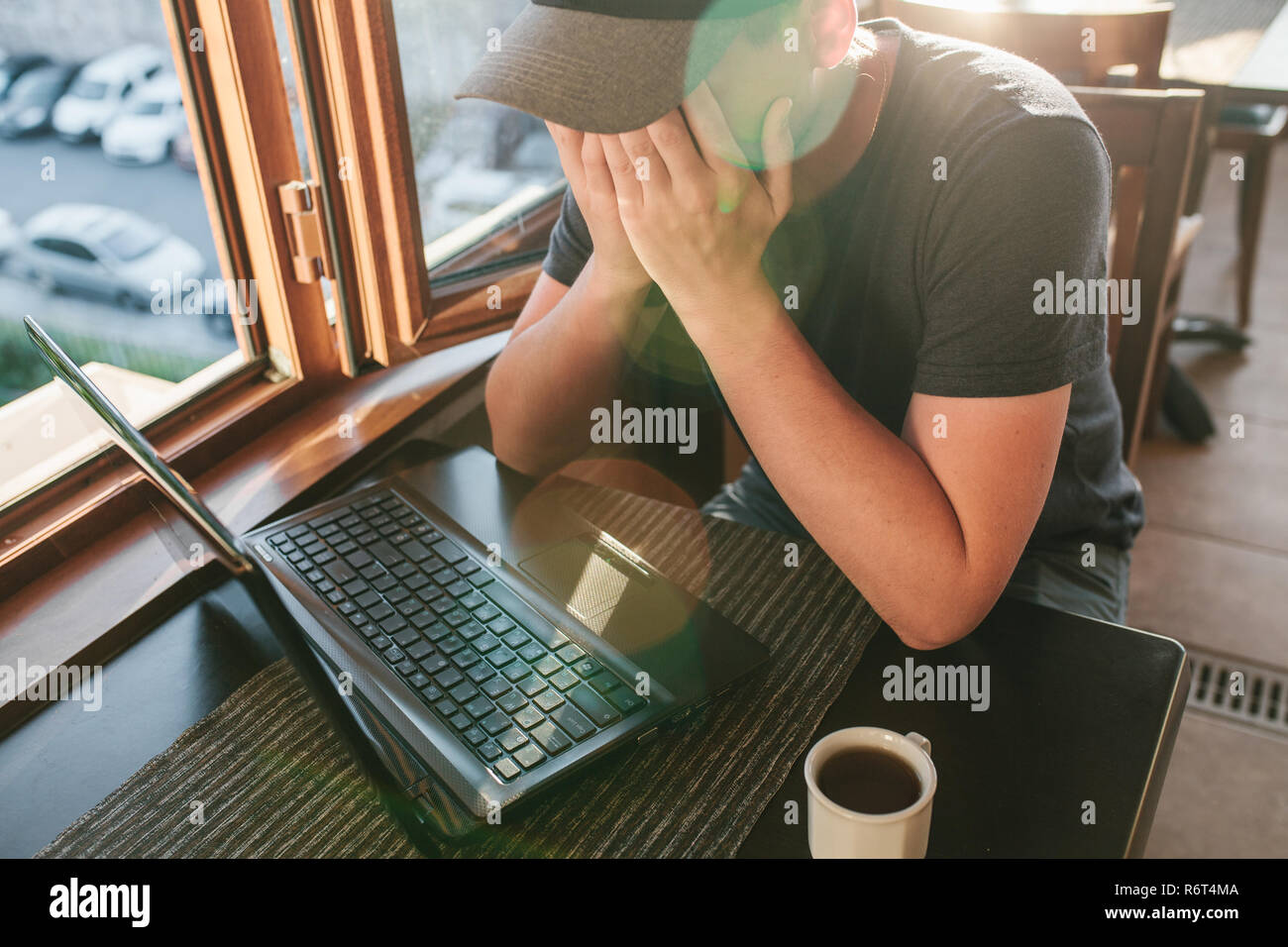 Man in cafe sits at table with computer near window and hidden face by hands Stock Photo