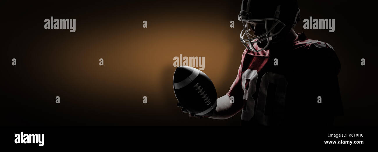 American football player standing with rugby ball and helmet against orange background with vignette Stock Photo