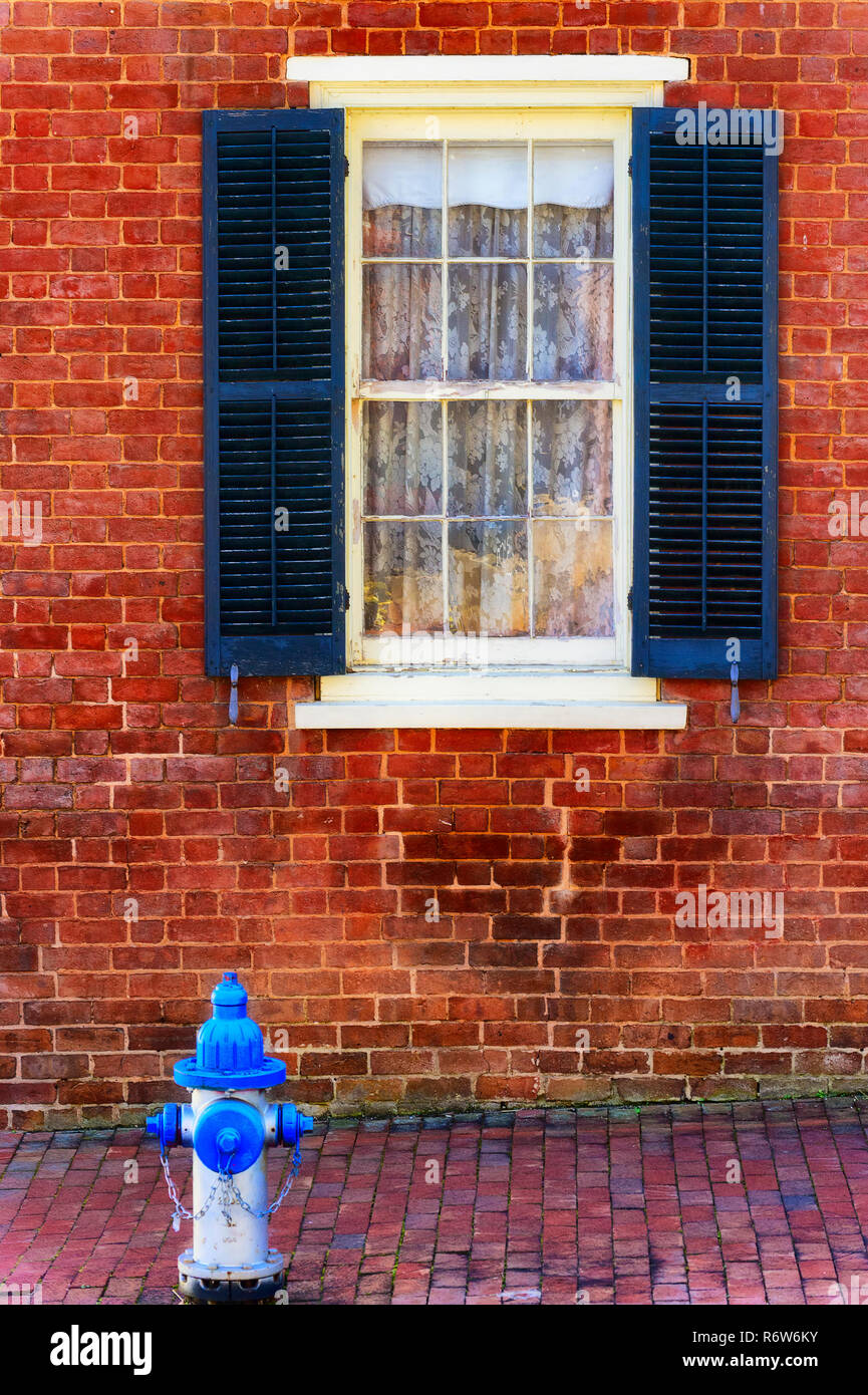 Brick sidewalk and exterior wall of an old building with a blue and white fire hydrant and a white window framed with dark blue wooden shutters. Stock Photo