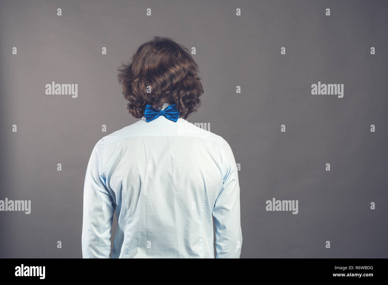 Back side view of a man with dark shaggy curly hair wearing blue shirt and bow tie against a grey background. Studio shot. Rear view people collection. Back view of person. Toned. Space for text. Stock Photo