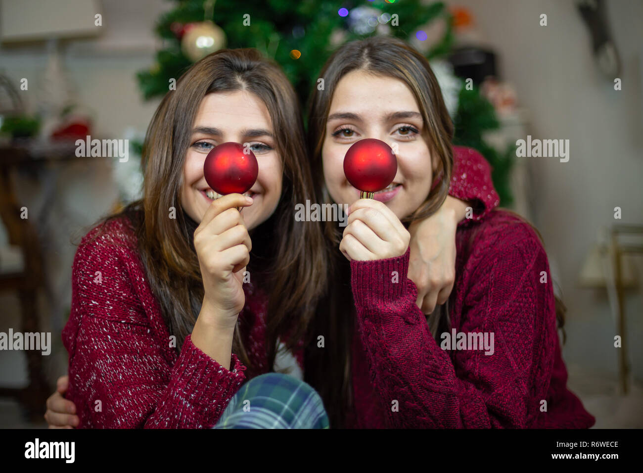 Content young women embracing while sitting on floor and making noses with red tree baubles Stock Photo