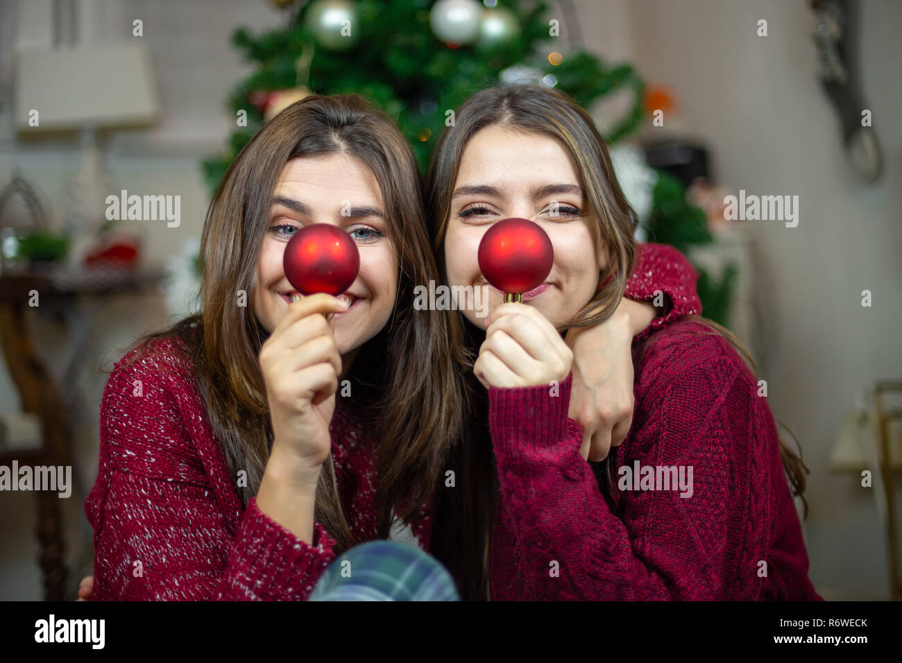Content young women embracing while sitting on floor and making noses with red tree baubles Stock Photo