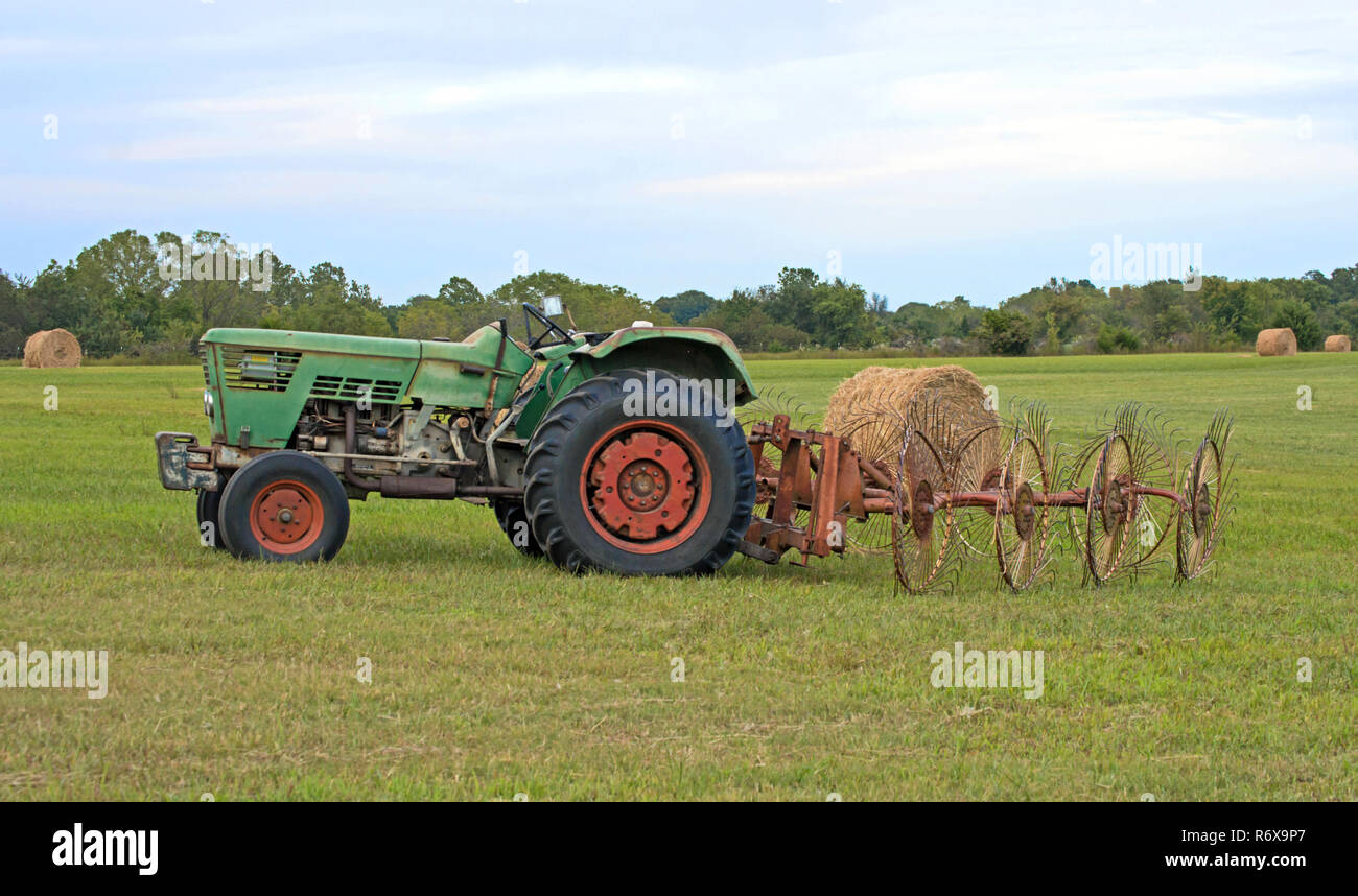 The old farm tractor sits idle in the field with hay rake attached.  The farmer has taken a brief break from baling hay on a hot summer day in Missour Stock Photo