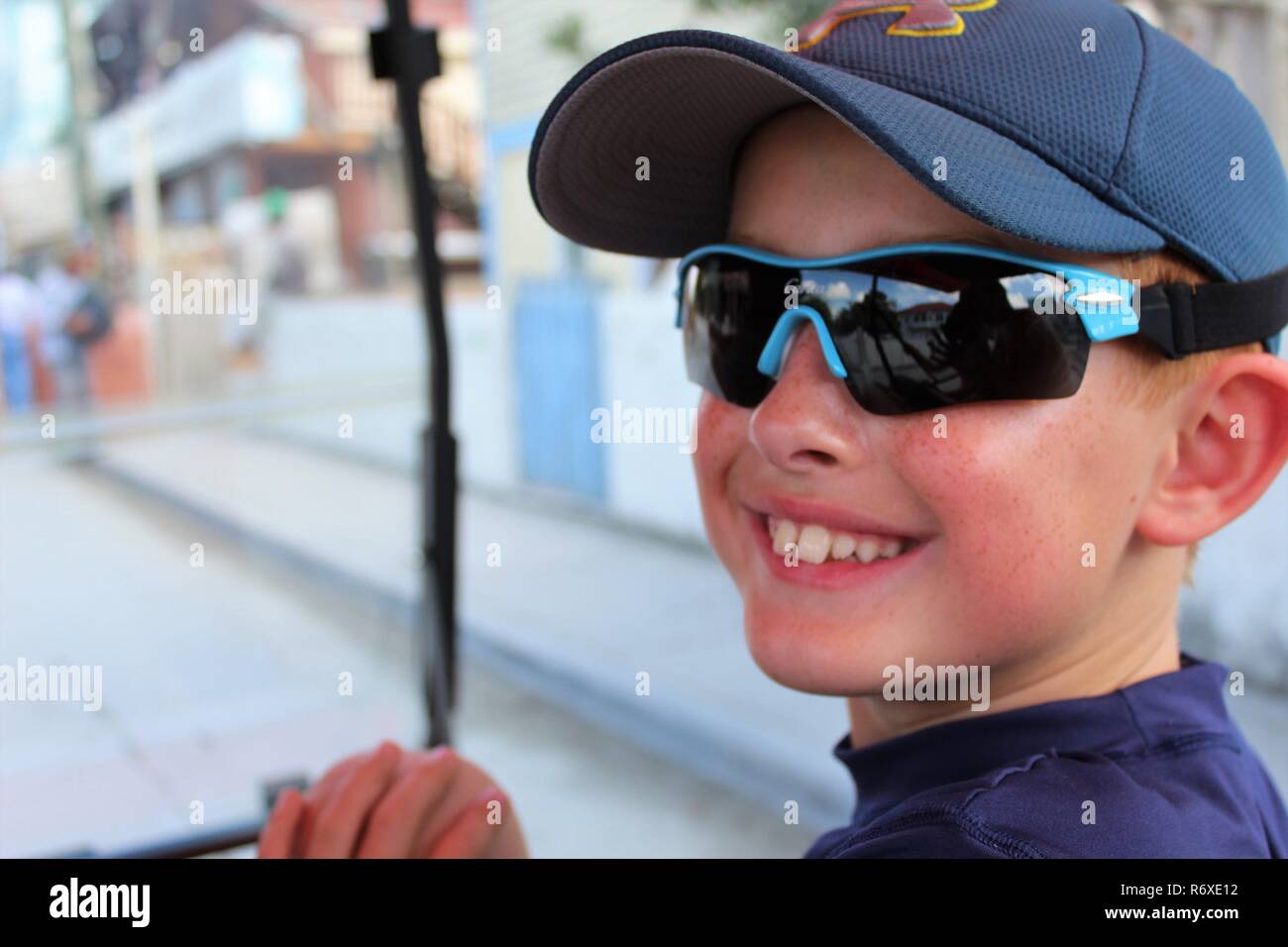 Cute red headed boy with sunglasses and hat turned to smile at camera while riding golf cart Stock Photo