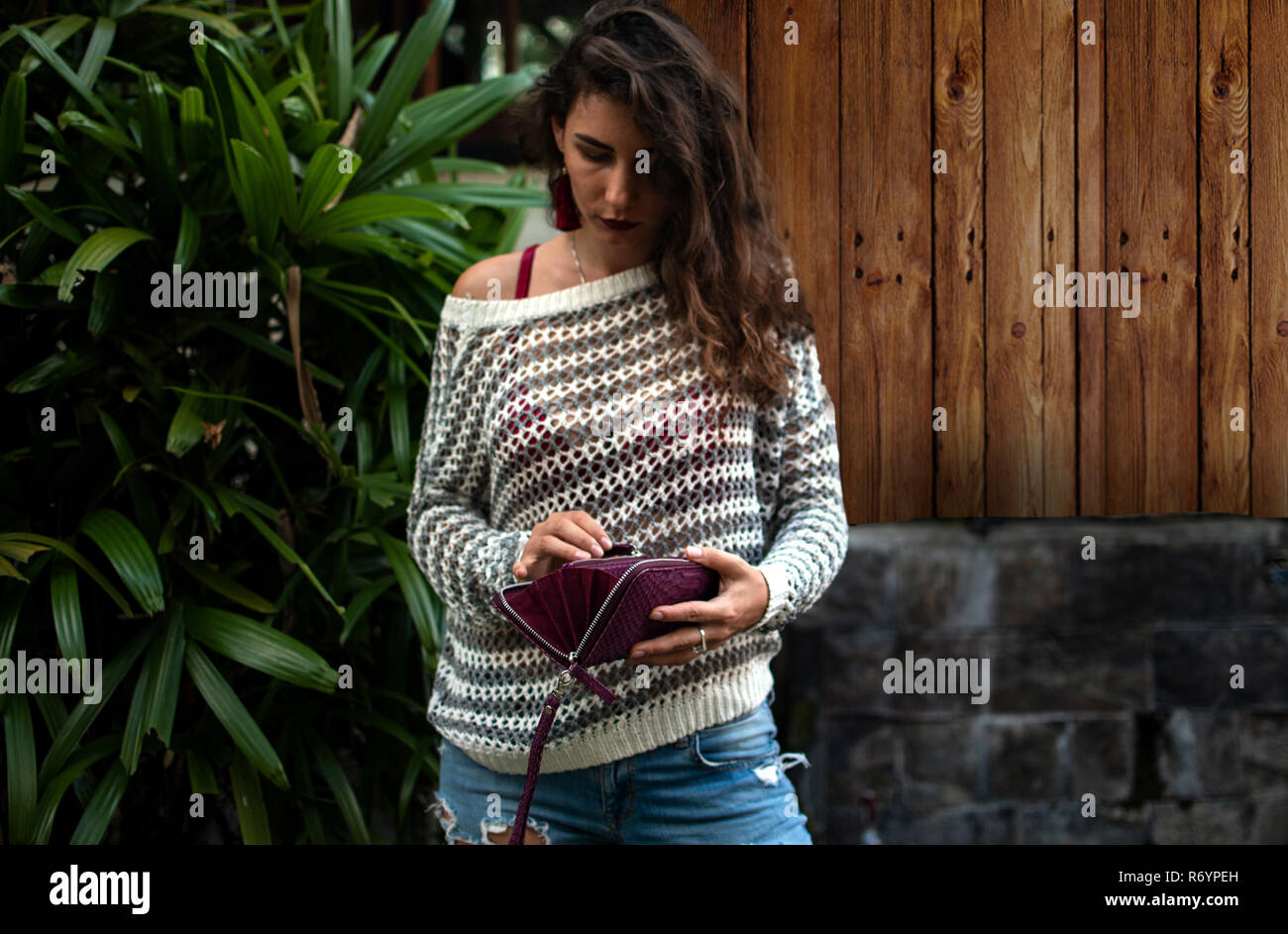 Woman holds a purple python purse in her hands. money and shoping. Woman's hands with wallet, nature background, palms leaves, wood, wearing jeans, fashion style Stock Photo