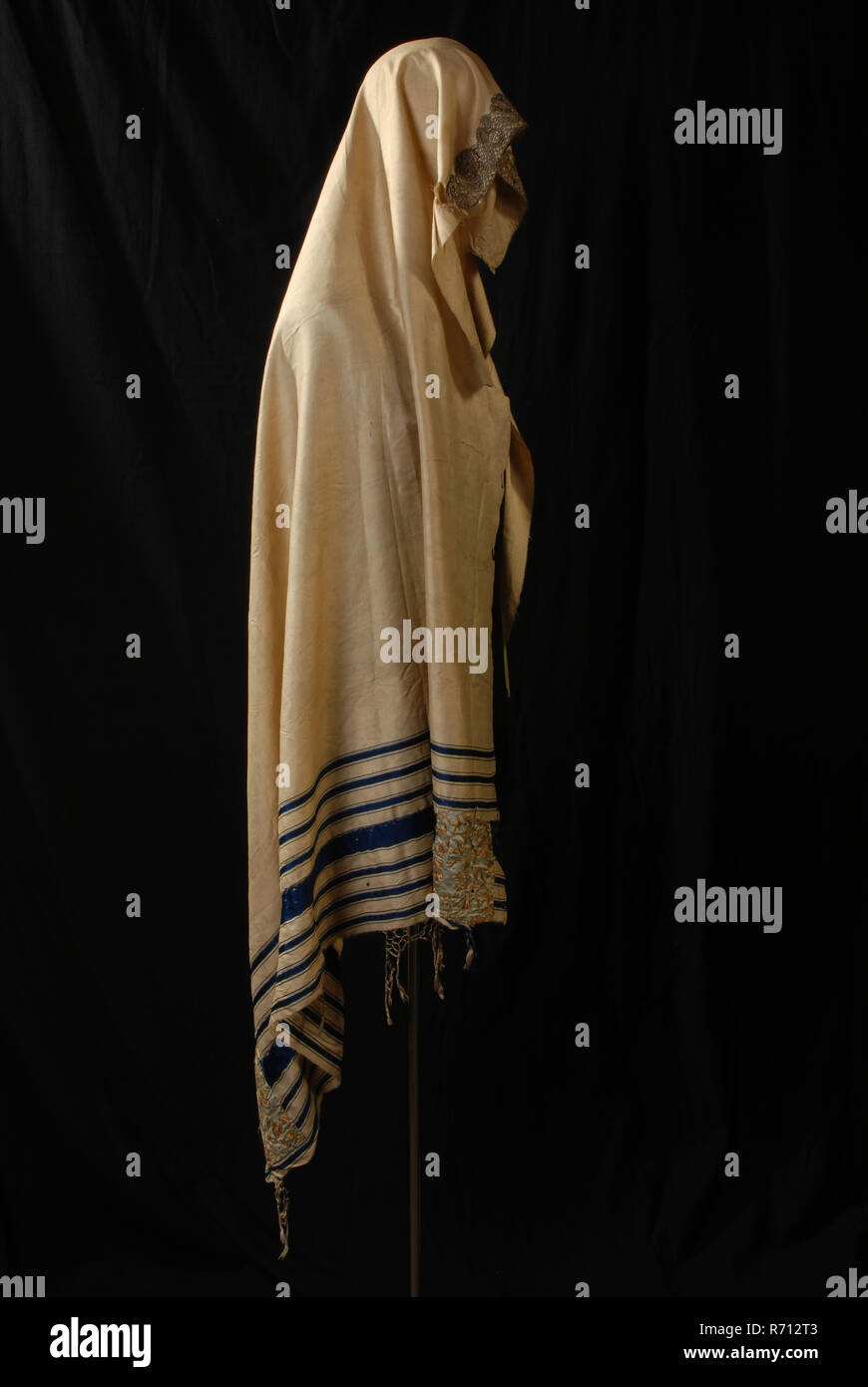 Jewish prayer scarf or tallit gadol of white silk with gold and silver embroidery, tallith liturgical clothing men's clothing apparel silk gold silver, textile appliqué White silk cloth with blue webs woven on one side and piece of silver brocade lace of about 35 cm long on the other side. Seven blue strips have been woven around and there are two rectangular pieces of fabric with gold embroidery. In these patches is hole for button cord to do. cardboard number is sewn on the strips. Along the short side are leftovers of frills made from the fraying of the fabric religion Judaism judaica praye Stock Photo