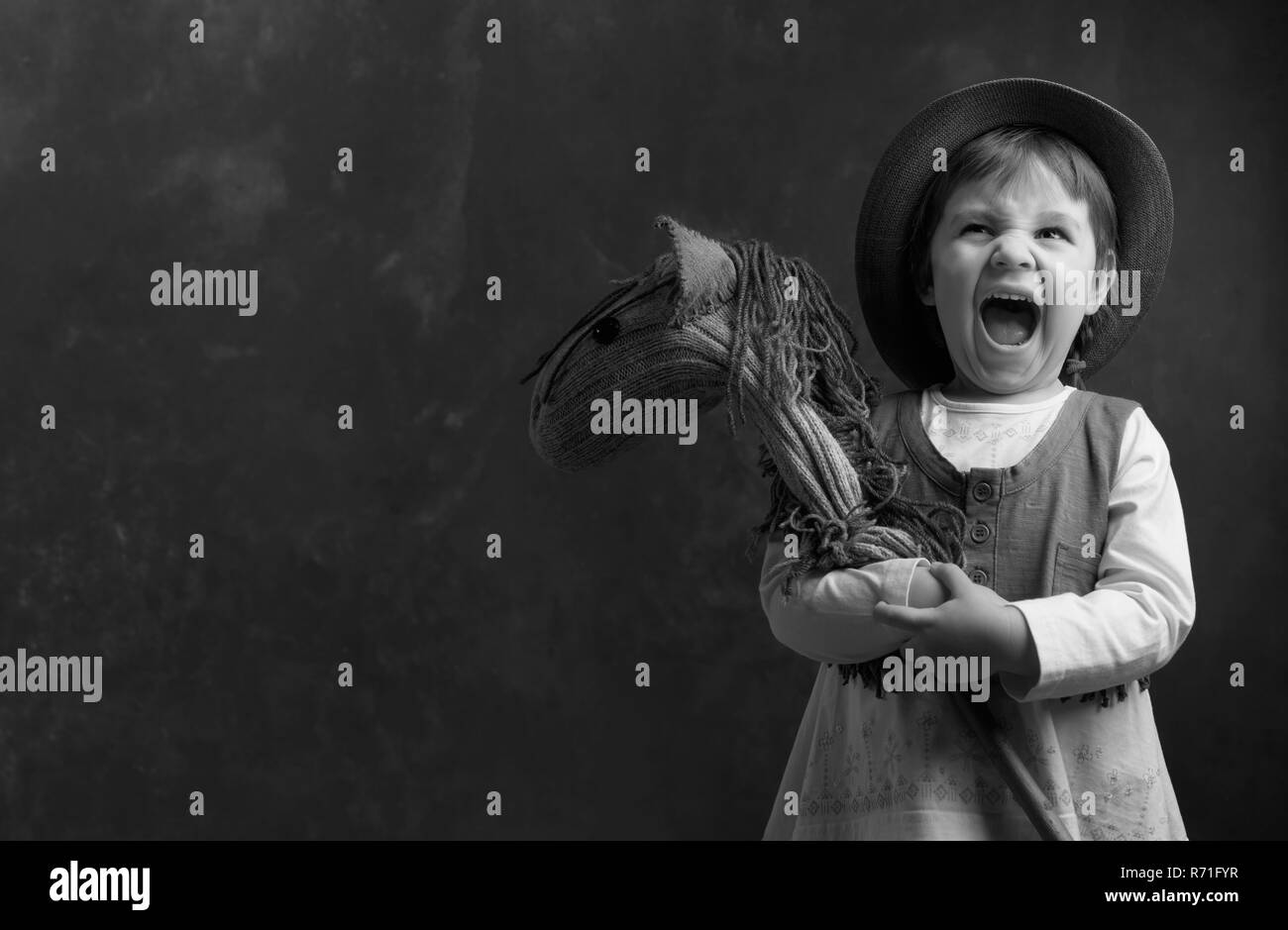 Screaming little girl with a homemade toy. Little girl dressed like a cowboy playing with a homemade horse. Expressive facial expressions. Copy space, Stock Photo