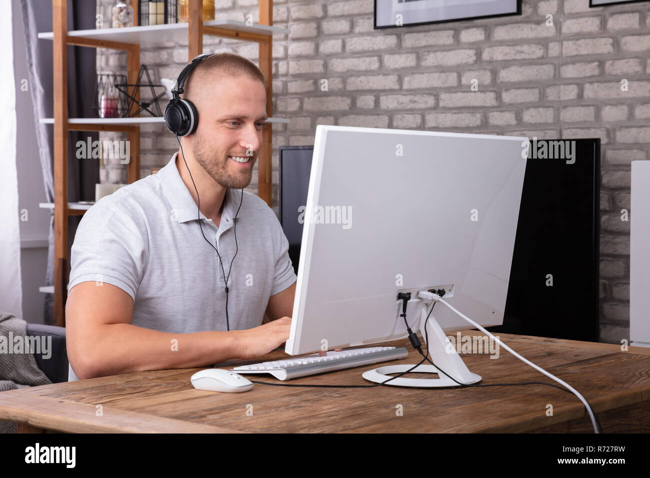 Smiling Young Man Using Headphone While Working On Computer Over Wooden Desk Stock Photo