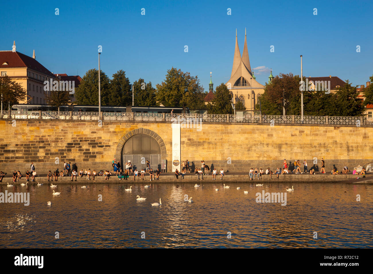 People relaxing on the embankment called Náplavka and watching swans in the river Vltava, behind are famous towers of Emmaus Monastery Stock Photo