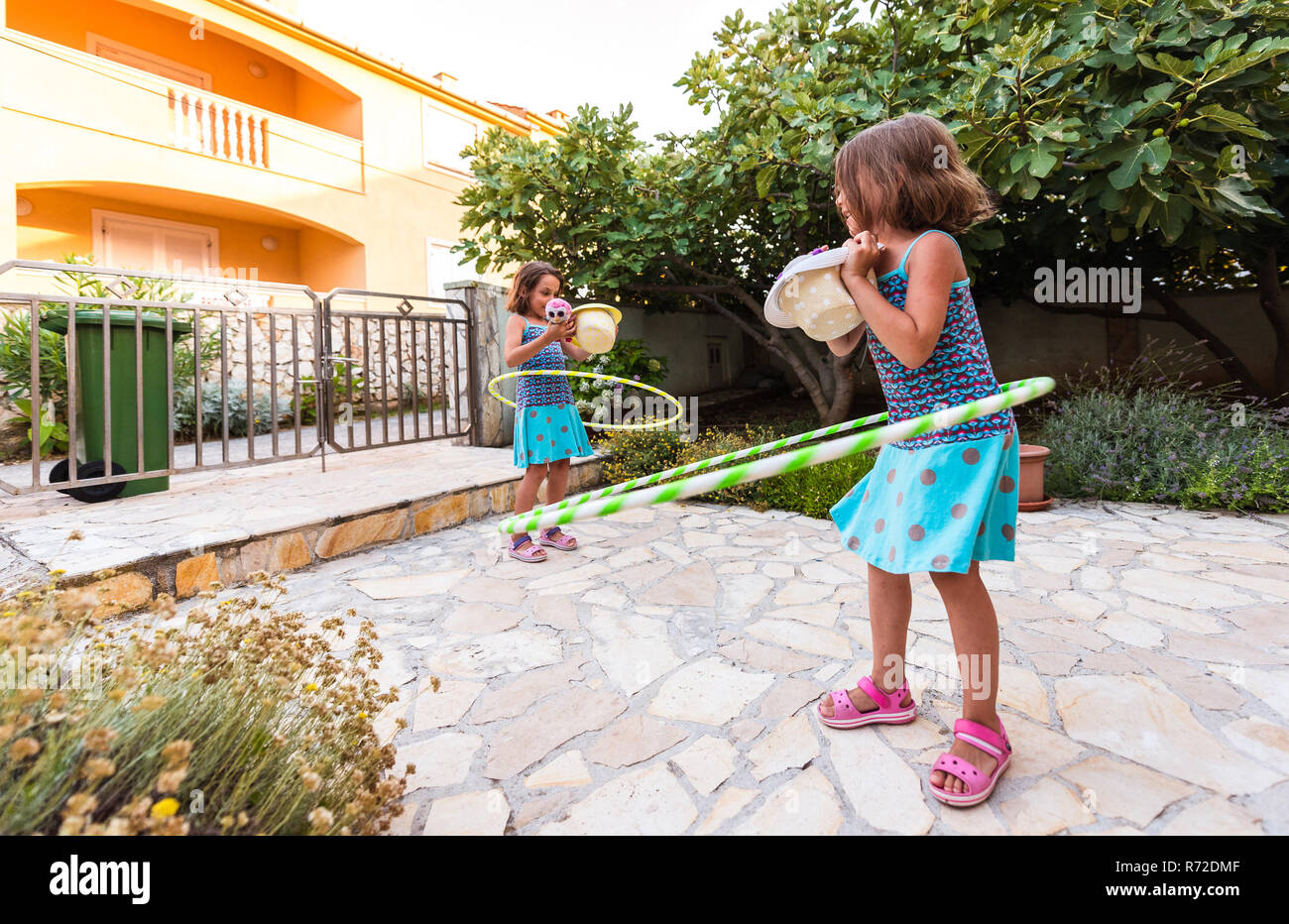 Identical twin sisters are playing on vacation with hula hoop. Sisters wearing blue dresses and hats are enjoying vacation hooping and laughing on sto Stock Photo
