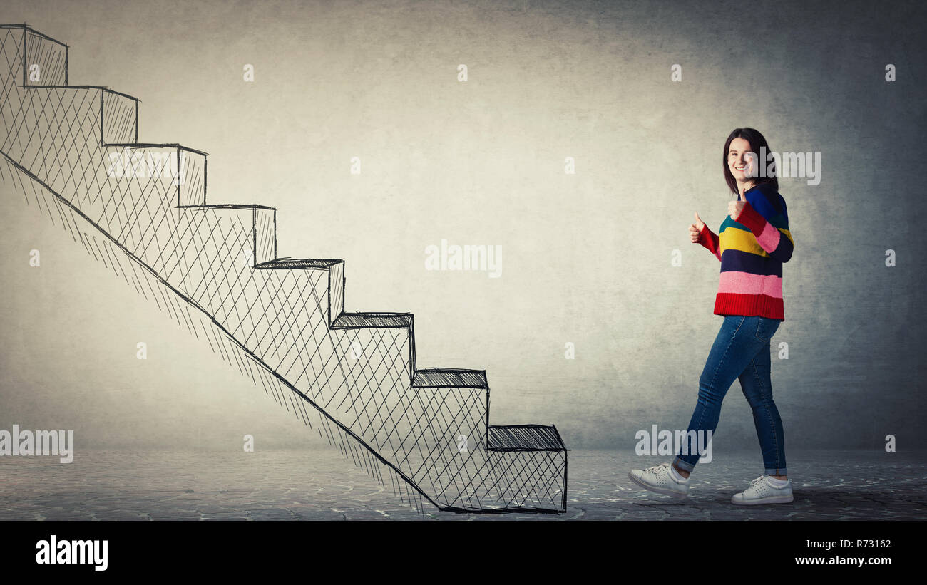 Full length side view portrait of young woman showing thumbs up gesture while stepping to a stairway. Going up as symbol of personal development and s Stock Photo