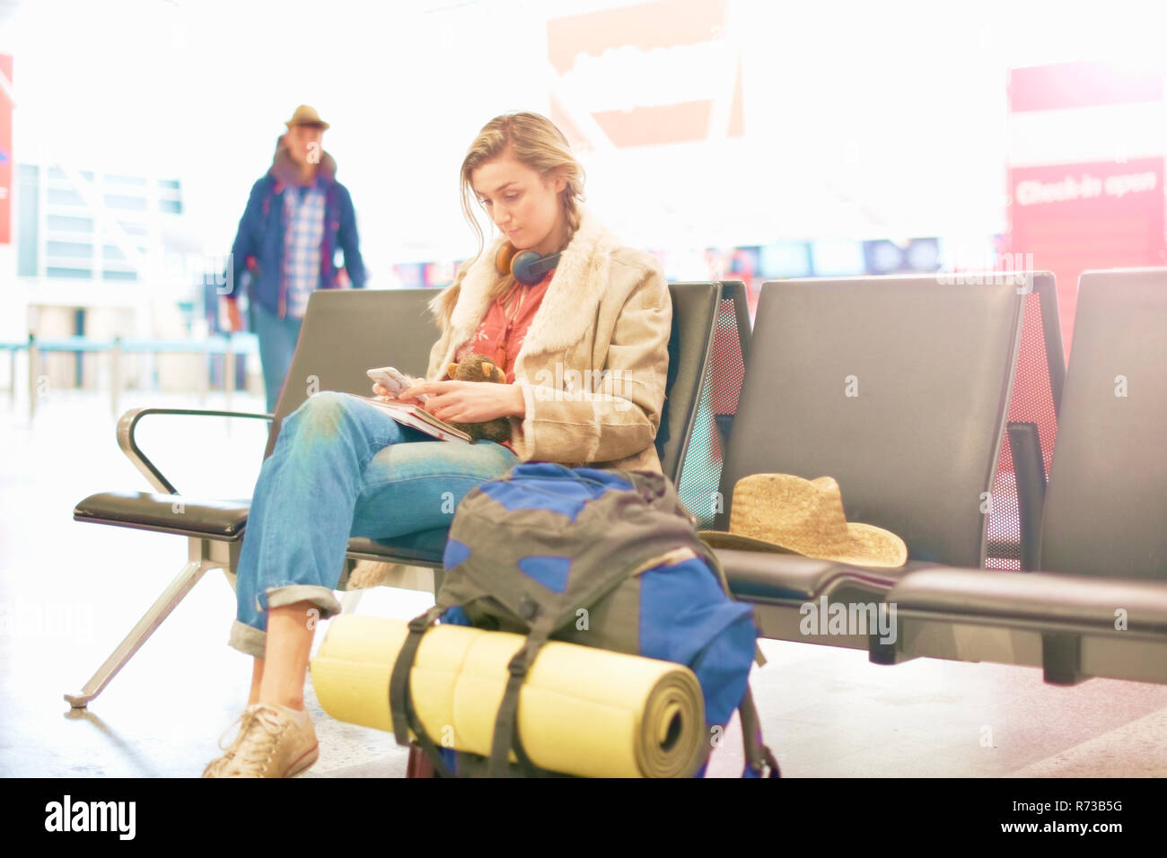Young woman at airport, sitting with backpack beside her, using smartphone Stock Photo
