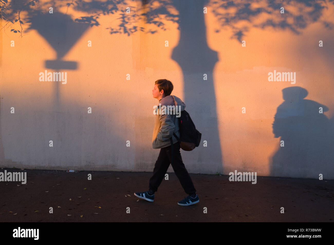 an eleven year old boy with backpack walks alongside a wall, covered with shadows, on his way to school early in the morning in Montpellier, France Stock Photo