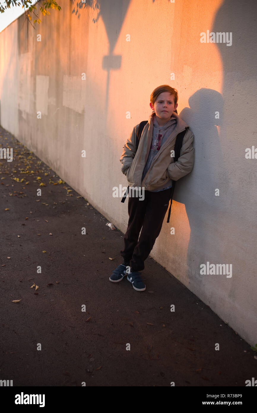 an eleven year old boy with backpack leans against a wall, covered with shadows, waiting for his school to open early in the morning in Montpellier Stock Photo