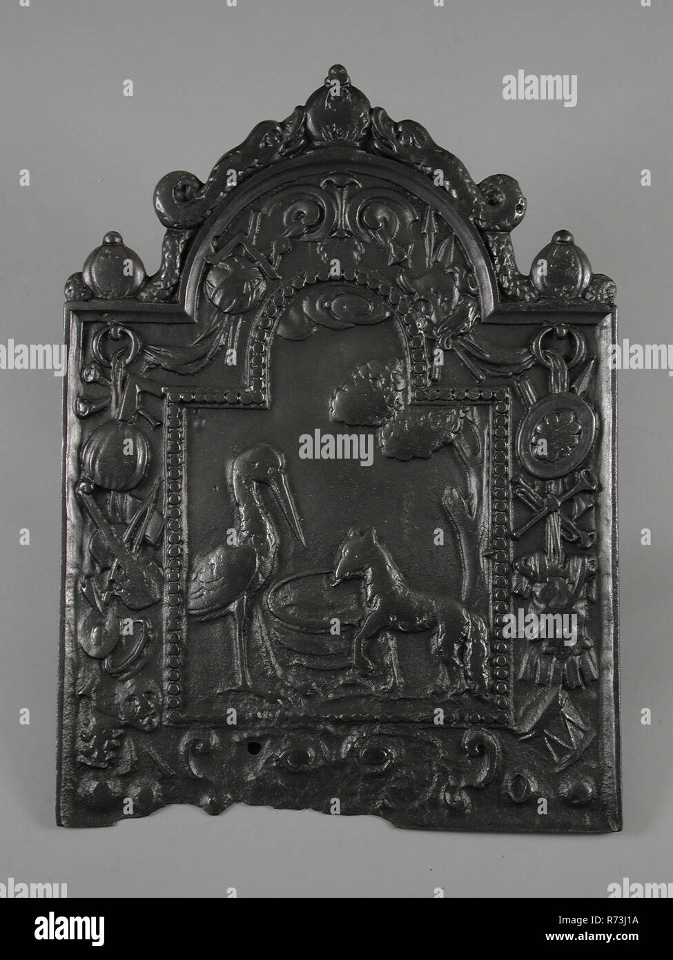 Fireback fable of fox and stork, after the fables of Jean de la Fontaine (1621-1695), fire place, cast Rectangular base with curved top with dolphins In the middle the first part of the fable of the fox and the stork Around wide border between the frame and the pearl necklace decorated with weaponry cuirass and various musical instruments living environment interior heating fable Jean de la Fontaine Stock Photo