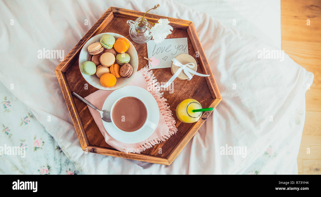 Breakfast in bed with i love you text on a note. Cup of coffee, juice, macaroons, rose and giftbox on wooden tray. Romantic breakfast in bed. Birthday Stock Photo