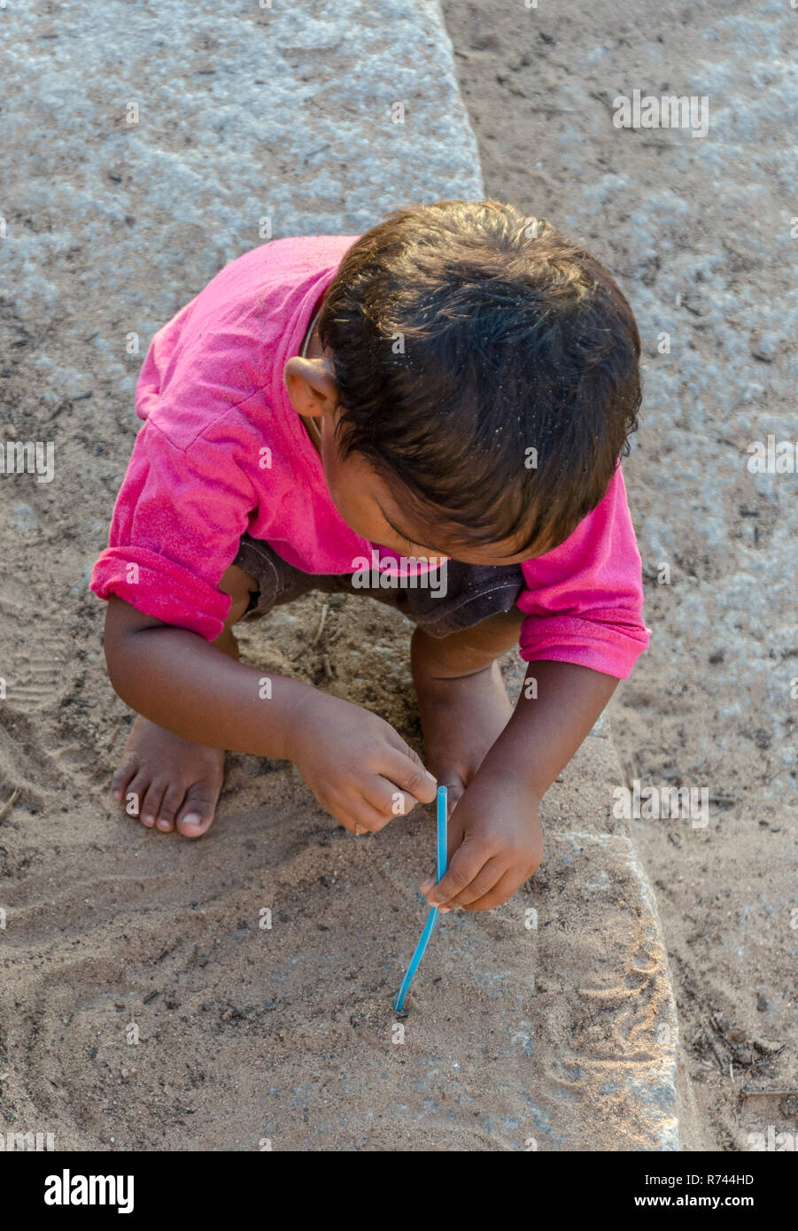 Overhead view of a young Indian kid playing with a plastic straw, pouring sand through it. Stock Photo