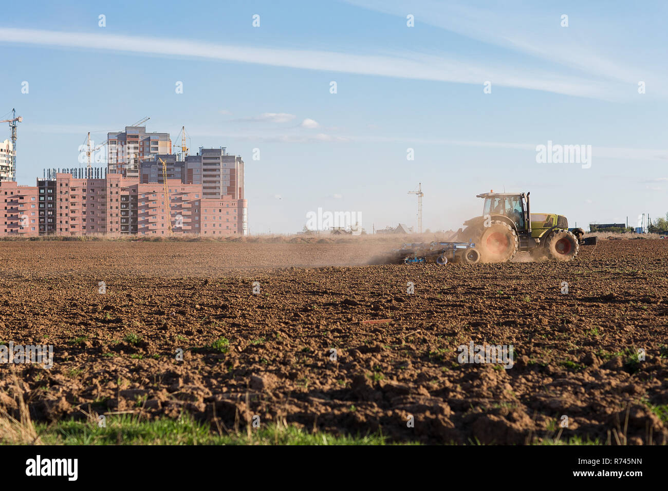 The industrialization of the vast expanses. Tractor plowing land on the background of houses under construction. Stock Photo