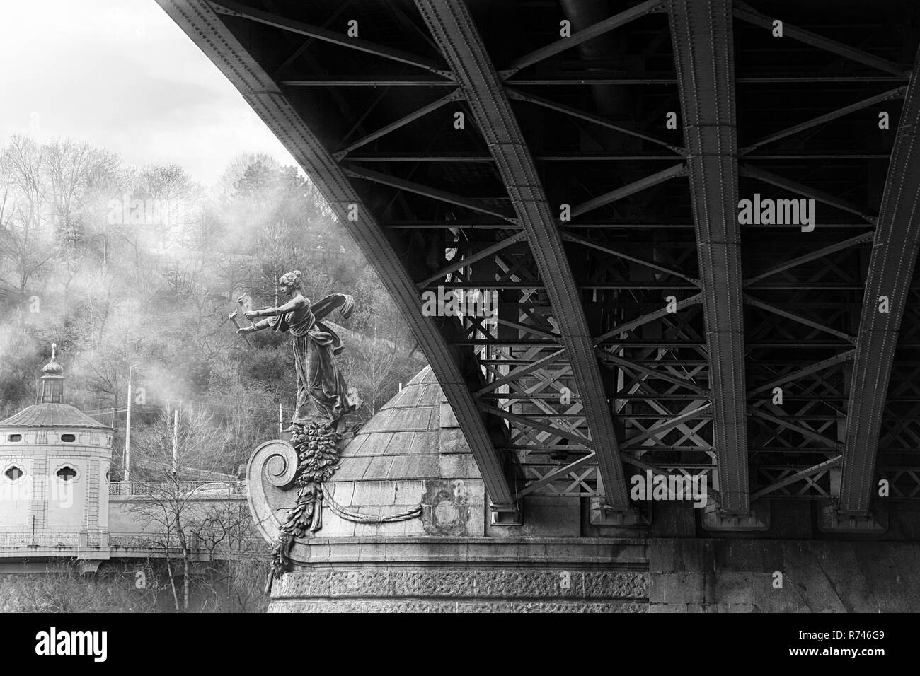 Foggy morning under the iron structure of a modern bridge, and a statue of a winged woman holding torches installed on the pillar. Black and white. Stock Photo