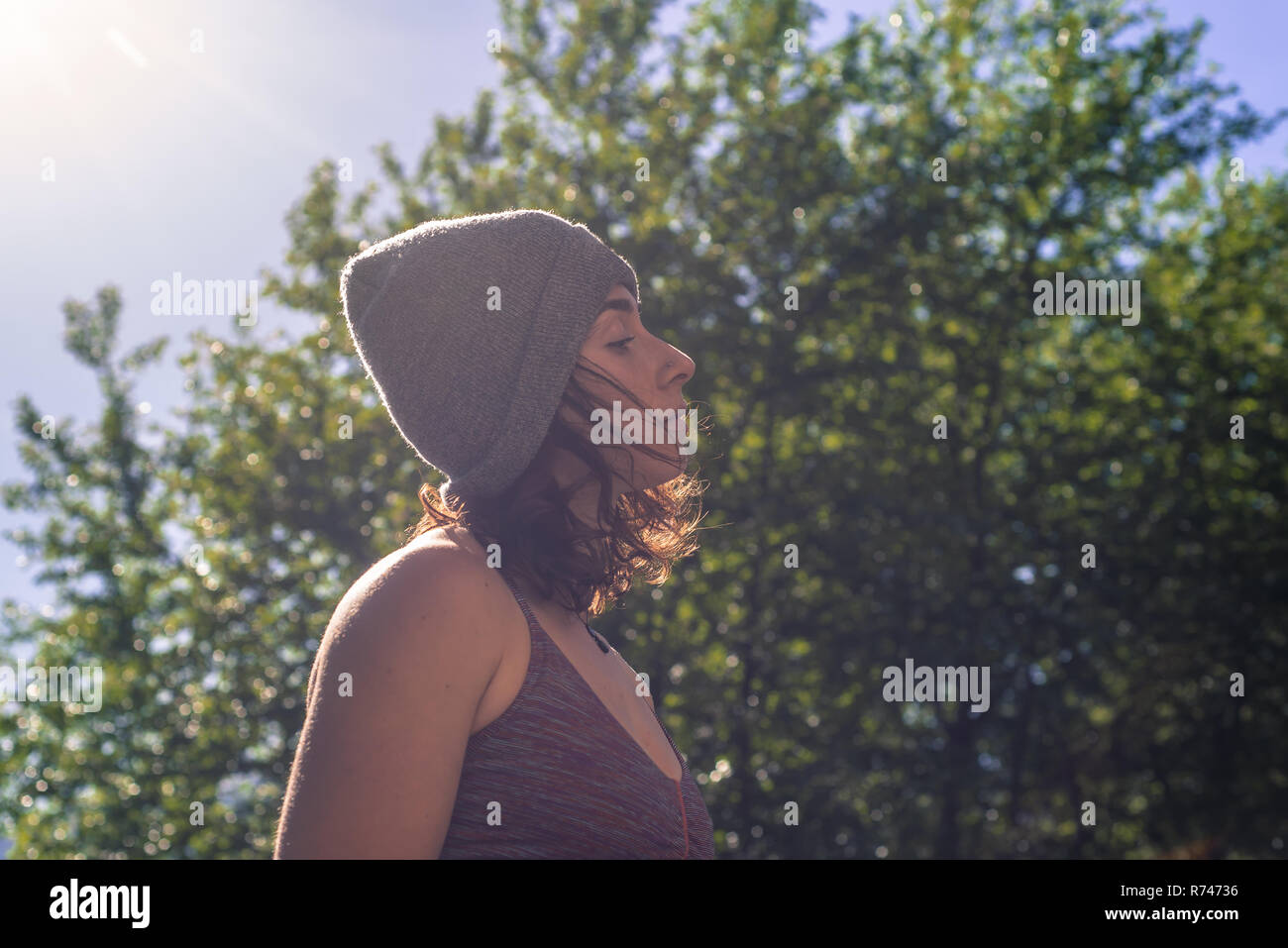 Young woman in knitted hat contemplating in front of tree foliage, backlit head and shoulder profile Stock Photo