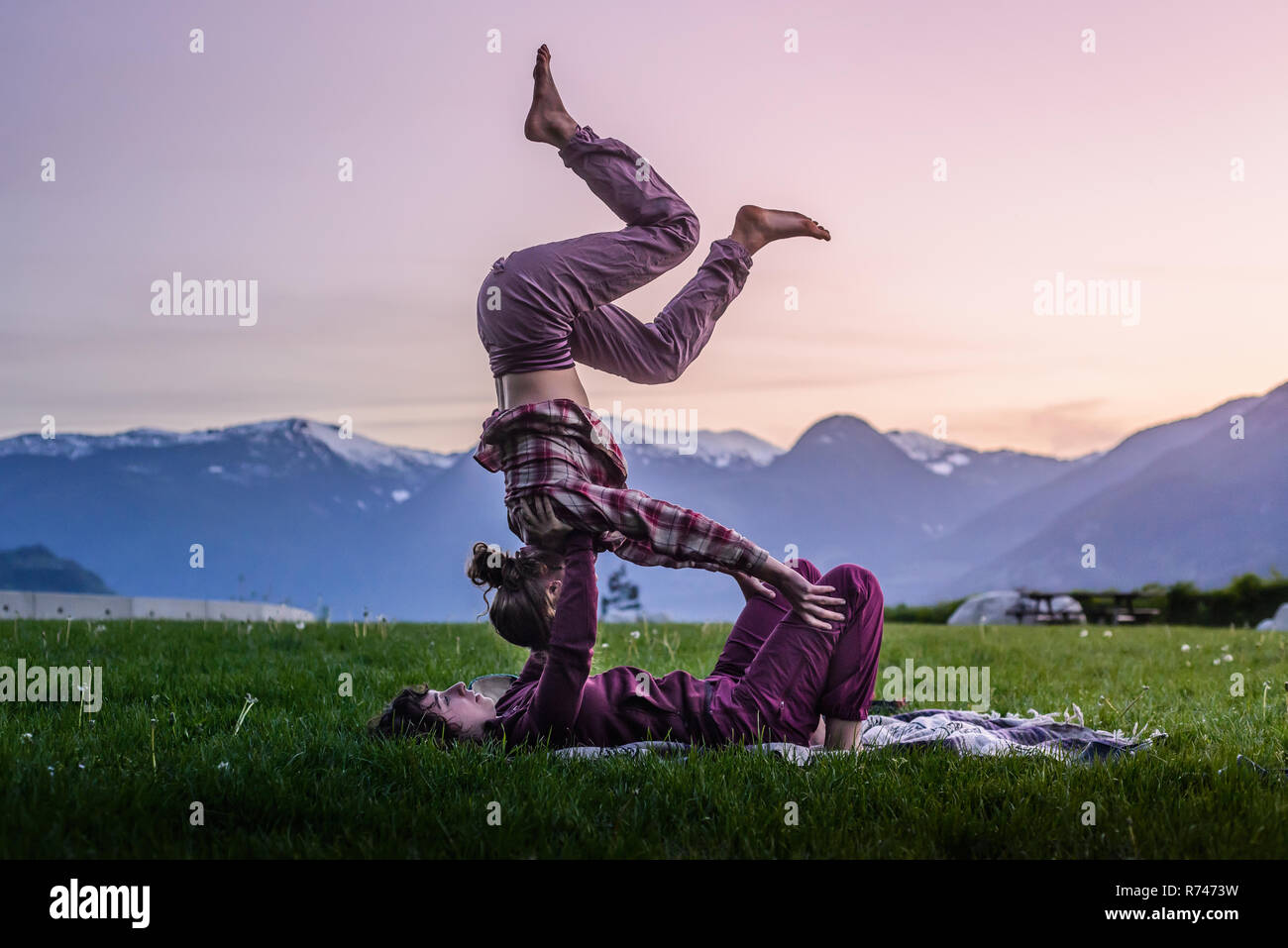 Two young women practicing acroyoga on grass in front of mountain range at sunset, Squamish, British Columbia, Canada Stock Photo