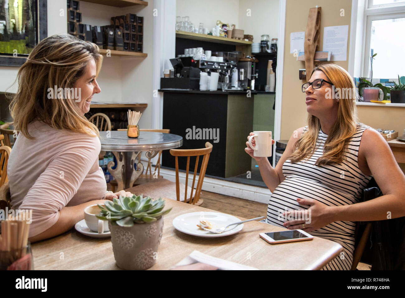 Pregnant mid adult woman and best friend chatting over coffee in cafe Stock Photo