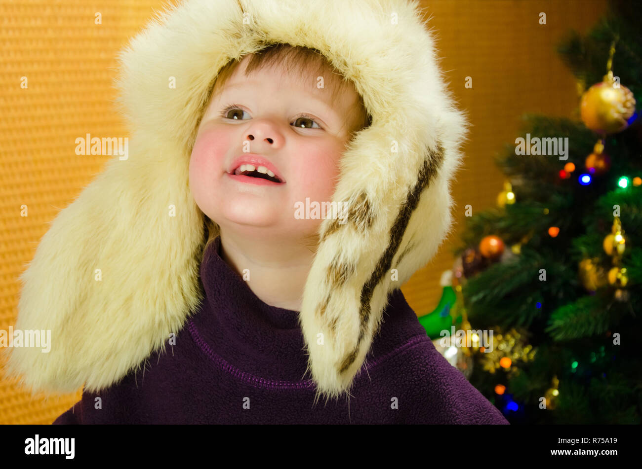 Portrait of two years old pretty little girl in white, russian, fur hat near a Christmas tree Stock Photo