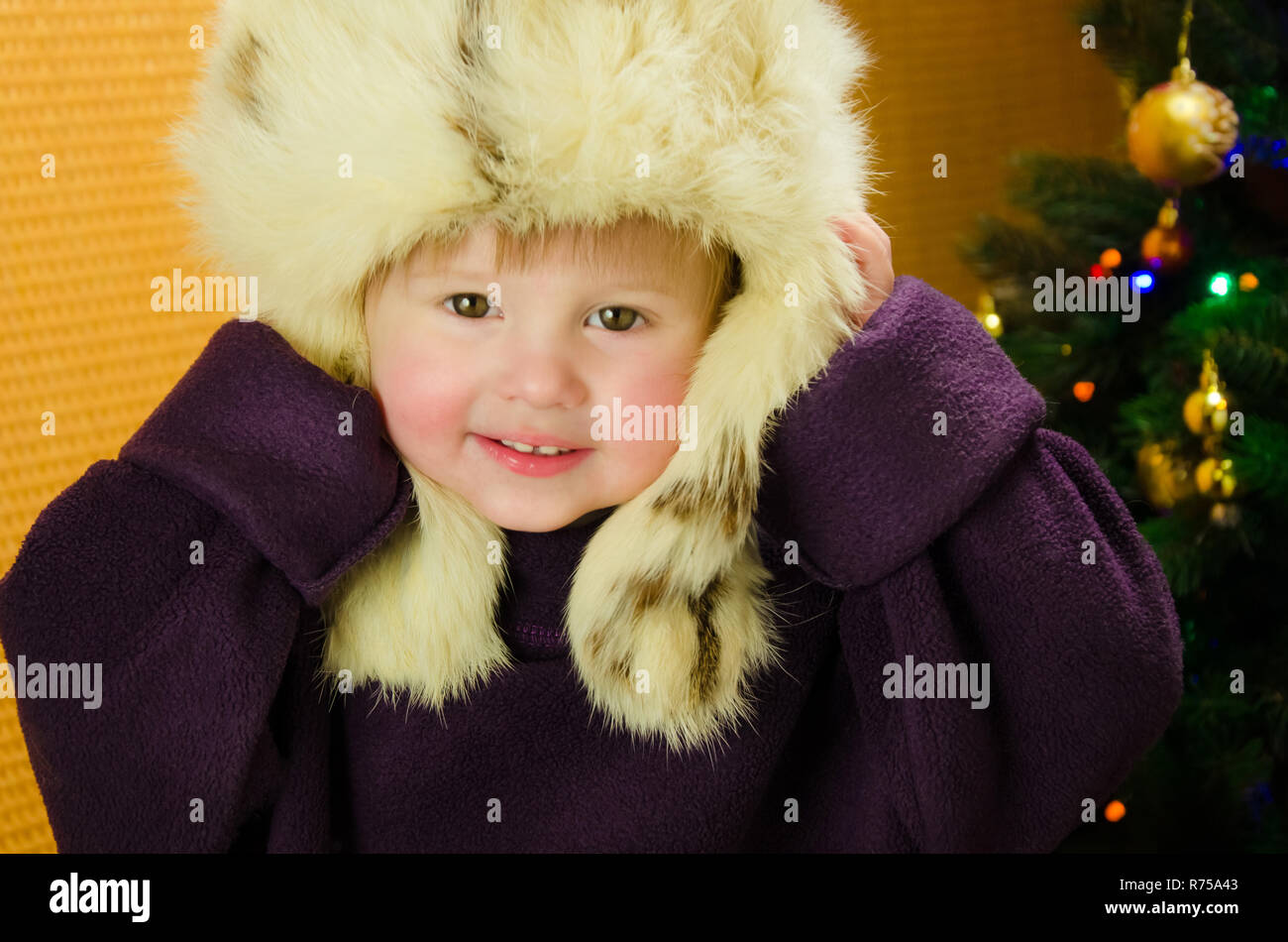 Portrait of two years old pretty little girl in white, russian, fur hat near a Christmas tree Stock Photo