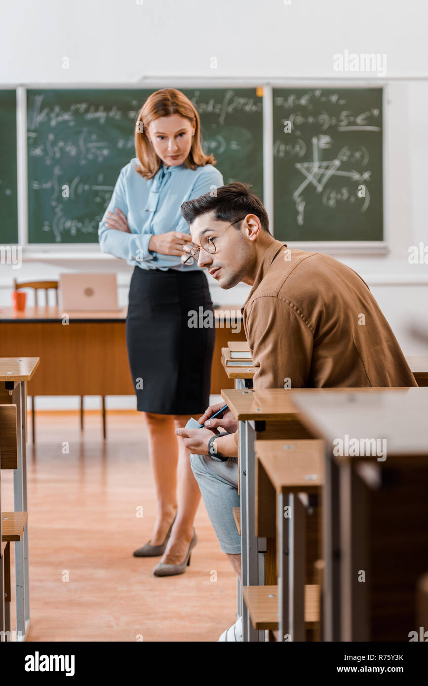 dissatisfied female teacher with arms crossed looking at male student cheating during exam in classroom Stock Photo