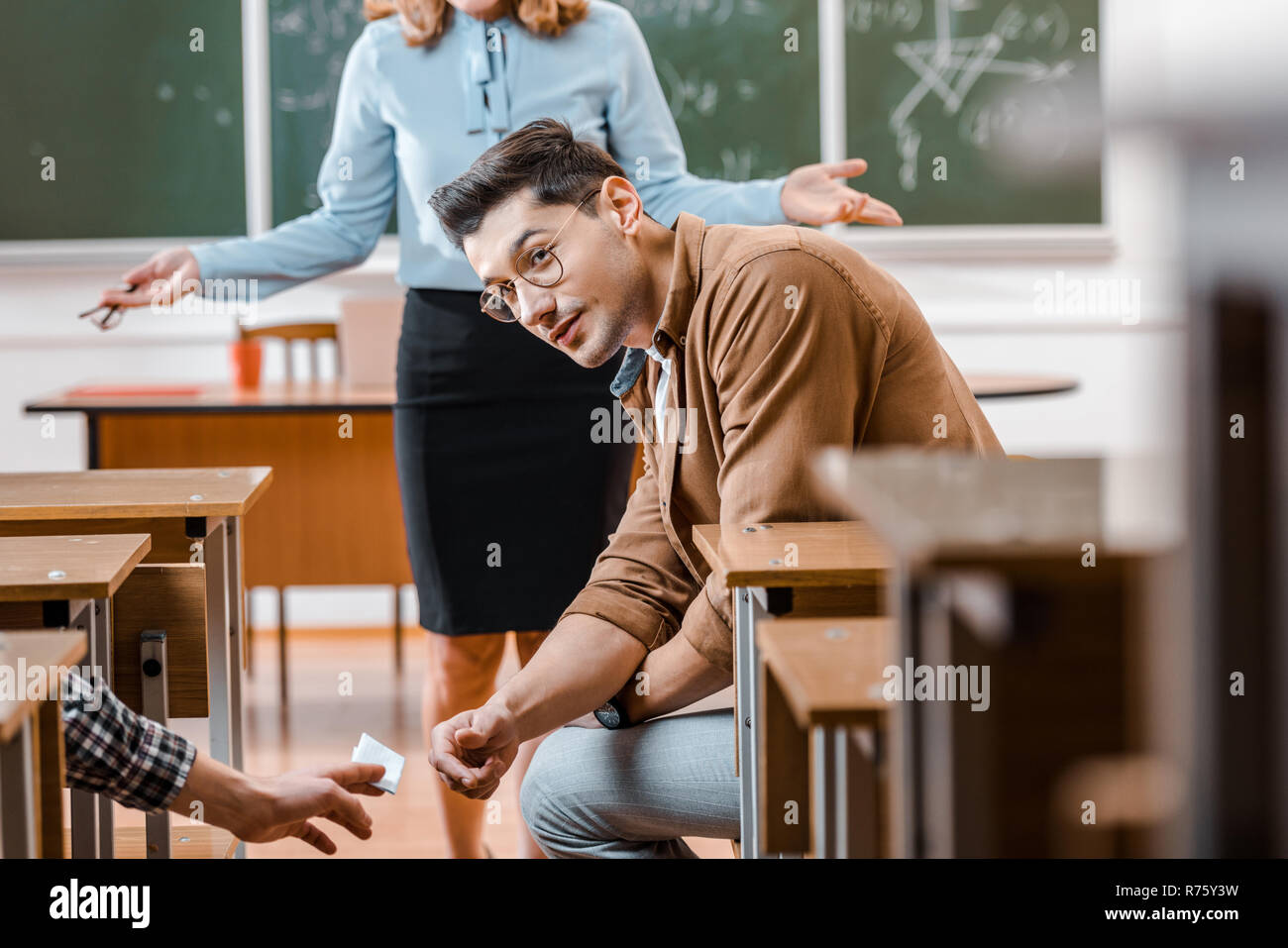 selective focus of dissatisfied female teacher looking at male student taking crib during exam in classroom Stock Photo