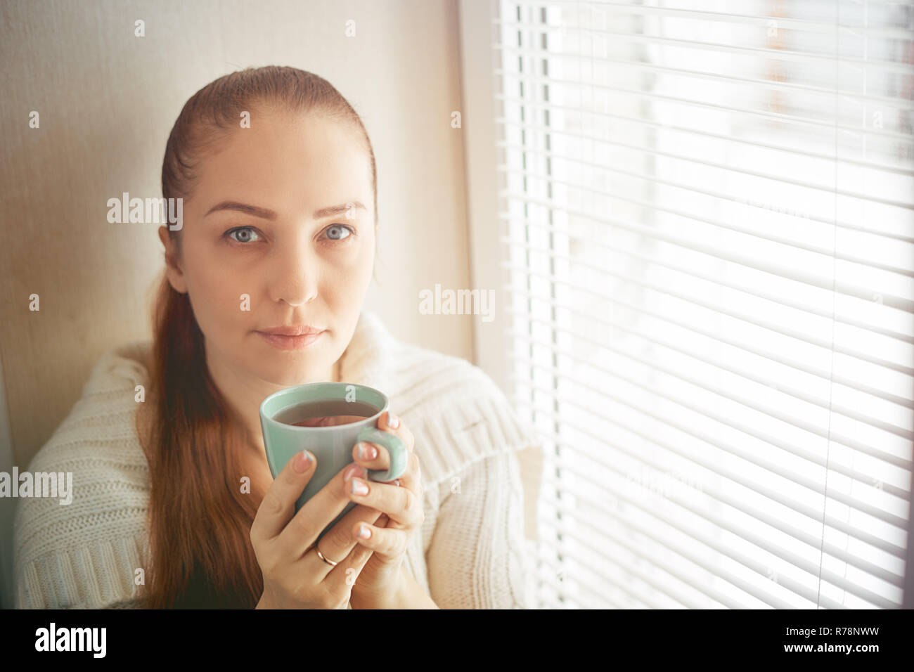 drinking tea sitting on the window of Caucasian beautiful middle-aged woman with red hair, toning Stock Photo
