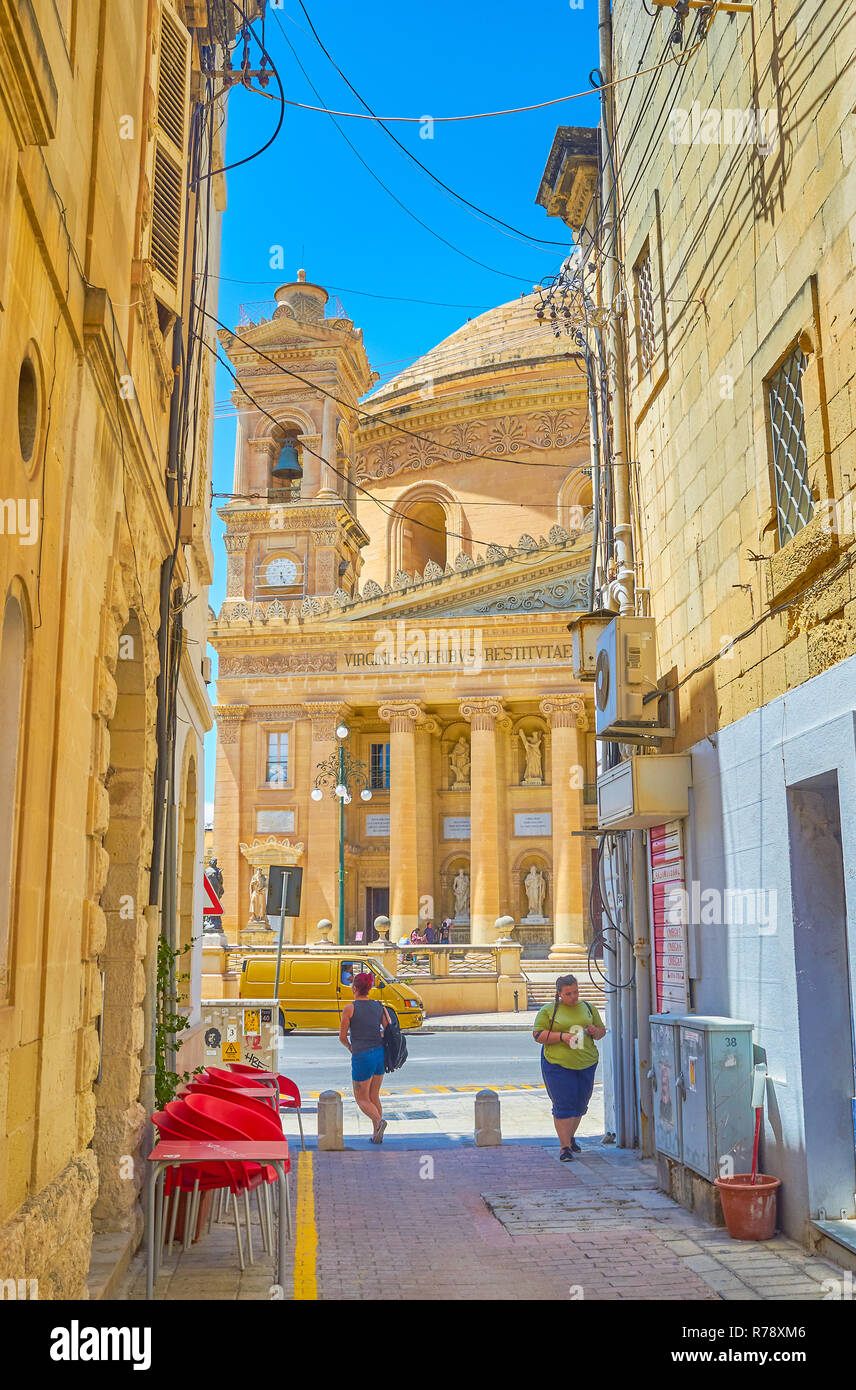 MOSTA, MALTA - JUNE 14, 2018: The view on the huge Basilica Rotunda through the narrow lane with small cafe, on June 14 in Mosta. Stock Photo