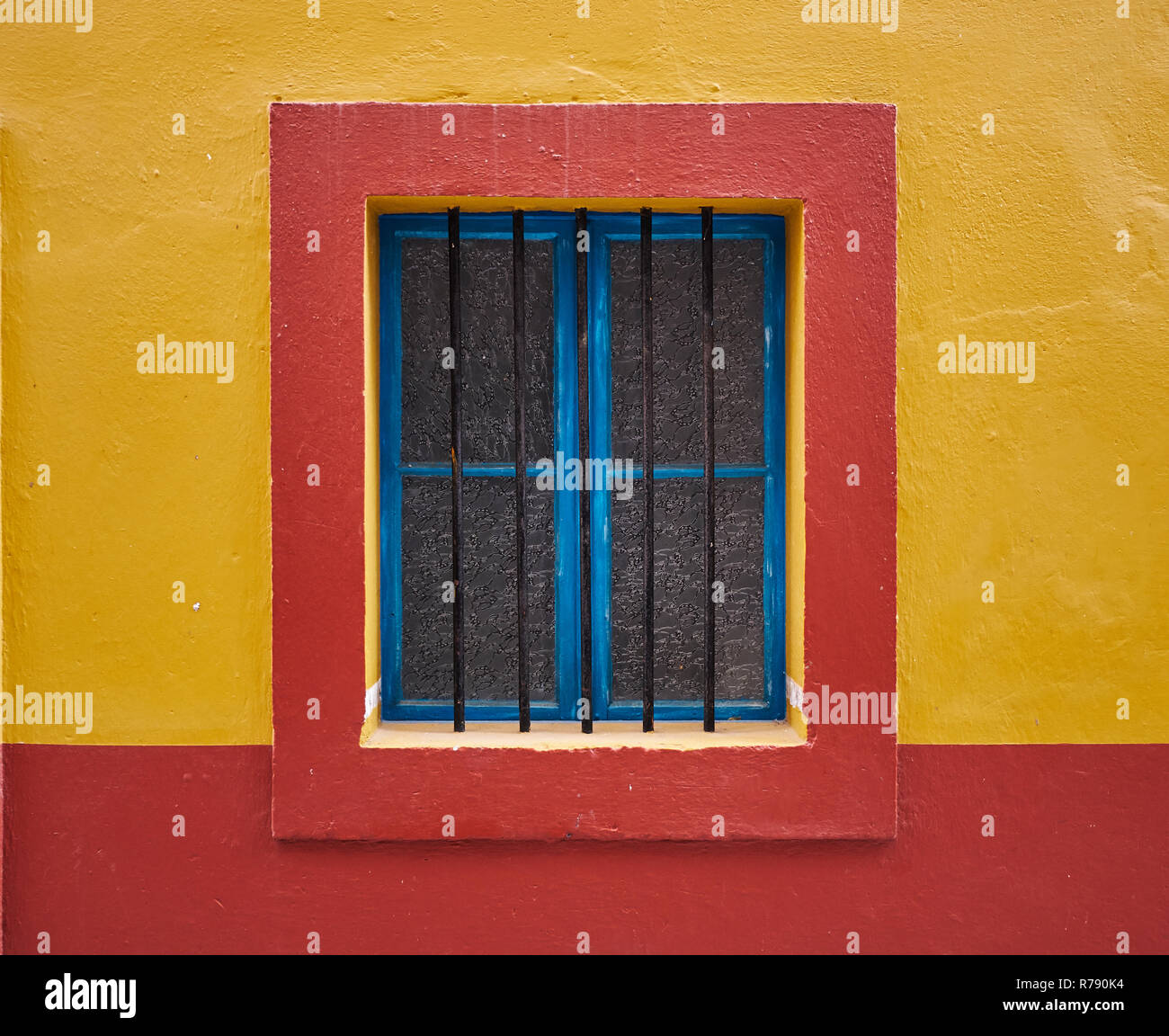 Minimalist wall image of a single centered blue window, with protective bars, framed in red on a yellow wall Stock Photo