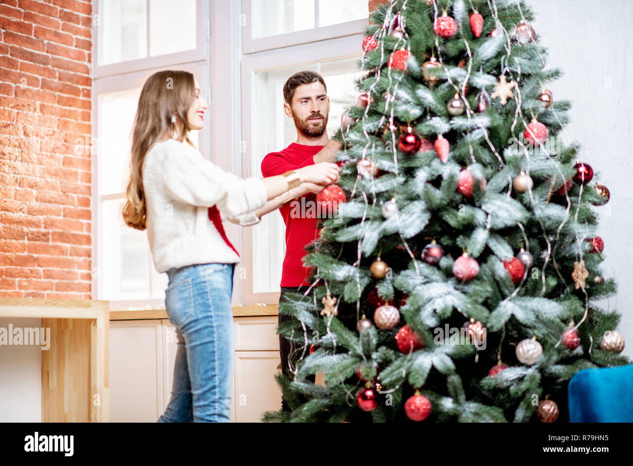 Young couple dressed in sweaters decorating Christmas tree preparing for a New Year holidays in their home Stock Photo