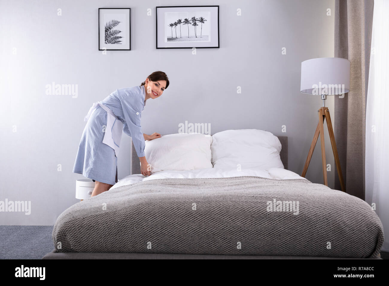 Smiling Female Housekeeper Making Bed In Hotel Room Stock Photo