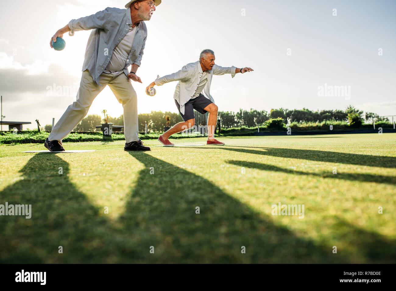 People playing a game of boules in a lawn with sun in the background. Two senior persons bending forward to throw  boules with their shadows on ground Stock Photo