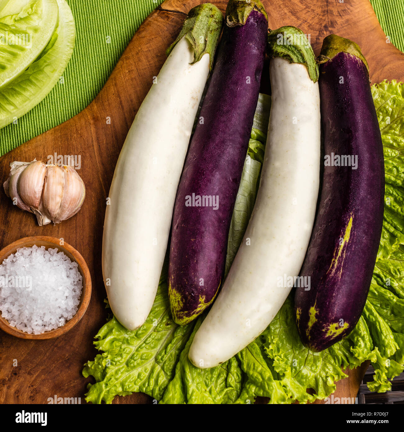 Fresh white and purple eggplant, lettuce and Romaine lettuce, chilly, cloves of garlic and wooden pot with sea salt on a wooden chopping board. Stock Photo