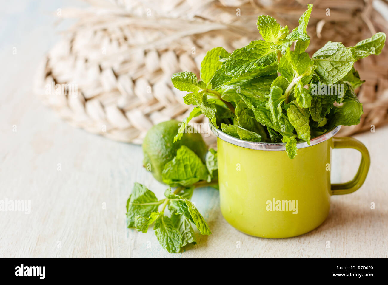 Fresh mint in a small lime green enameled cup on a wooden white and blue washed surface. Straw hat and a lime on the background. With space Stock Photo