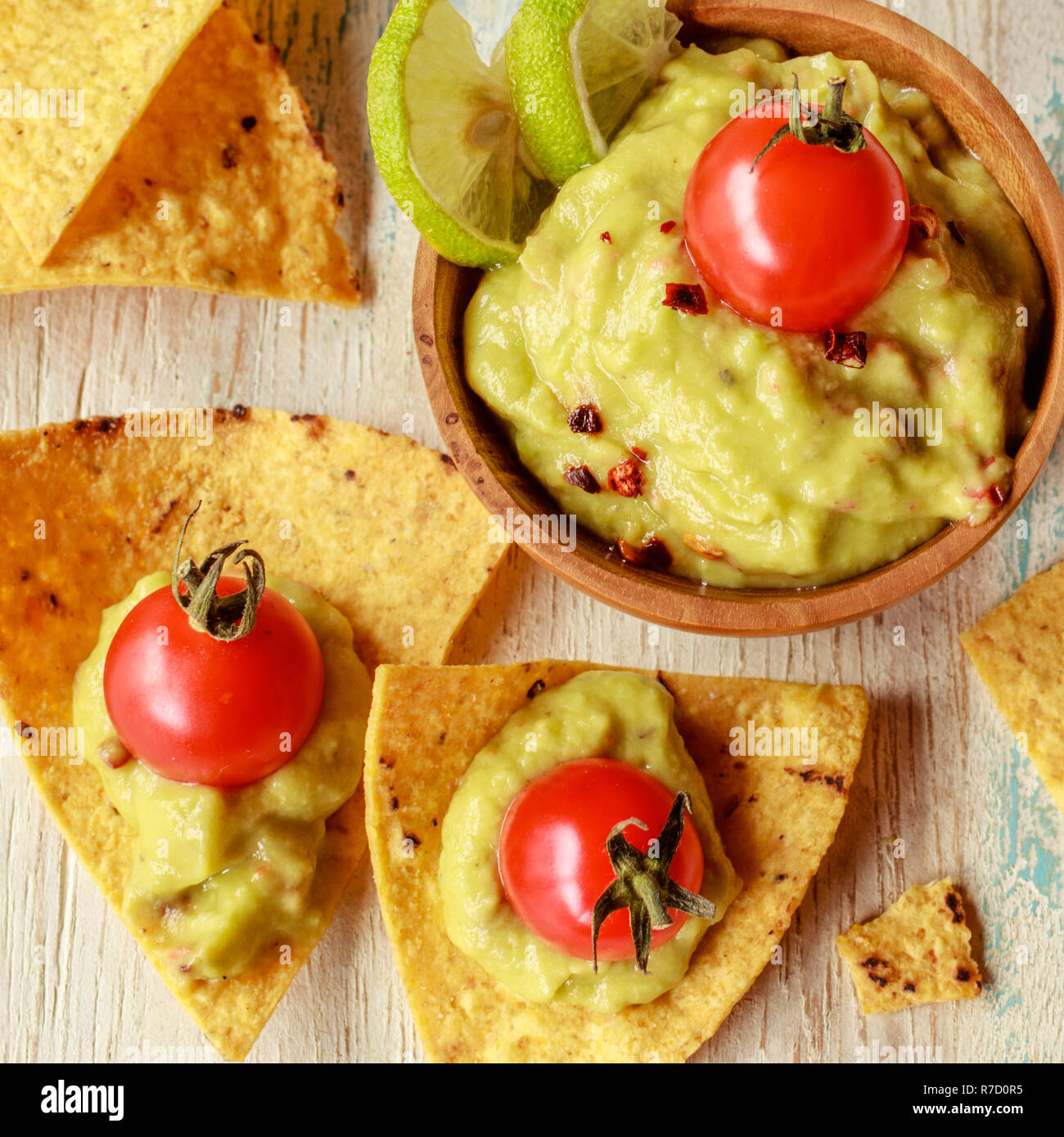 Corn chips nachos and guacamole in a wooden bowl decorated with cherry tomatoes and lime slices on a wooden white washed table. Stock Photo