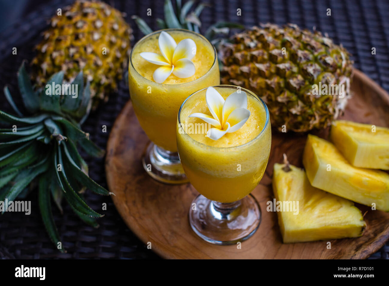 Two glasses of fresh pineapple juice decorated with Plumeria flowers, two whole pineapples and tree big cut pieces on a round wooden tray. Stock Photo