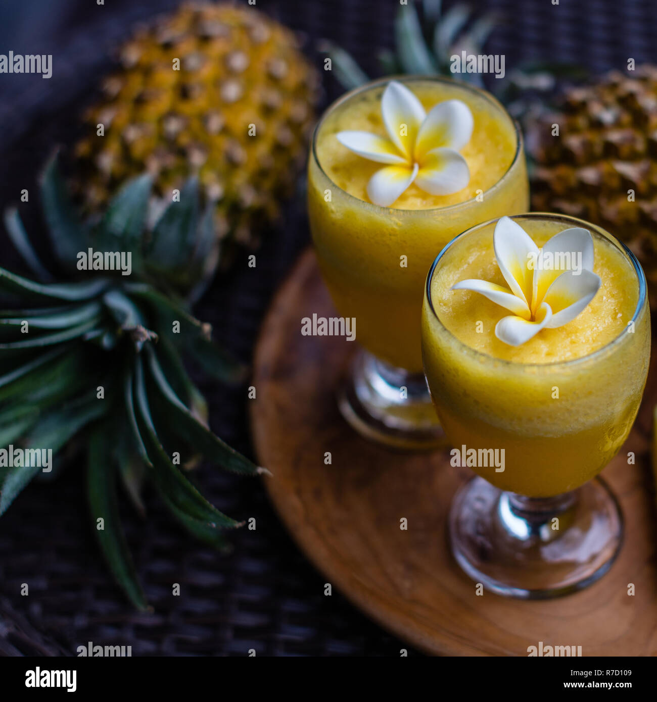 Two glasses of fresh pineapple juice decorated with Plumeria flowers, two whole pineapples and tree big cut pieces on a round wooden tray. Stock Photo