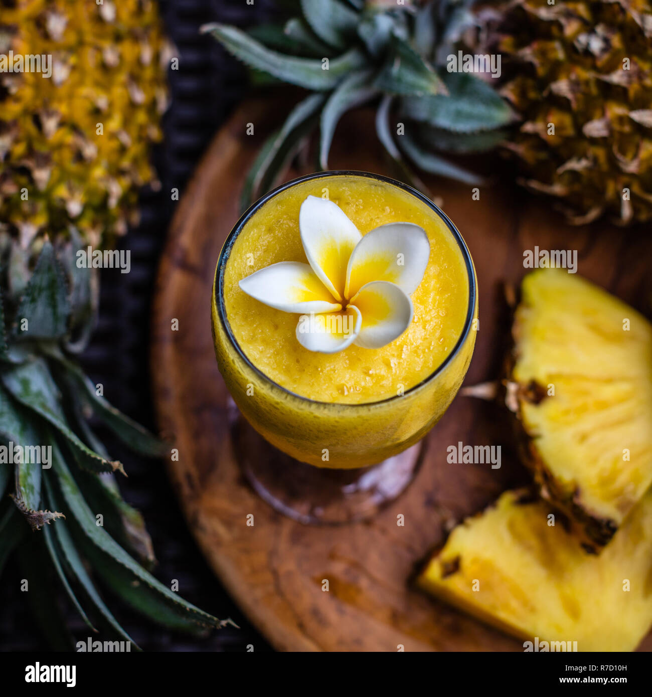 A glass of fresh pineapple juice decorated with Plumeria flower, two whole pineapples and tree big cut pieces on a round wooden tray. Stock Photo