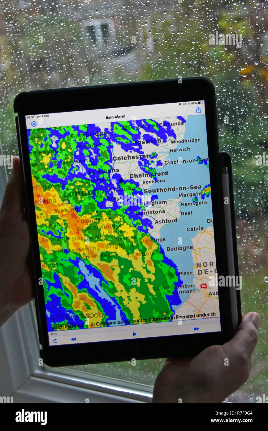 Woman hands holding ipad pro tablet rain alarm app uses real time colour radar data superimposing rainfall on local map wet weather on window Essex UK Stock Photo